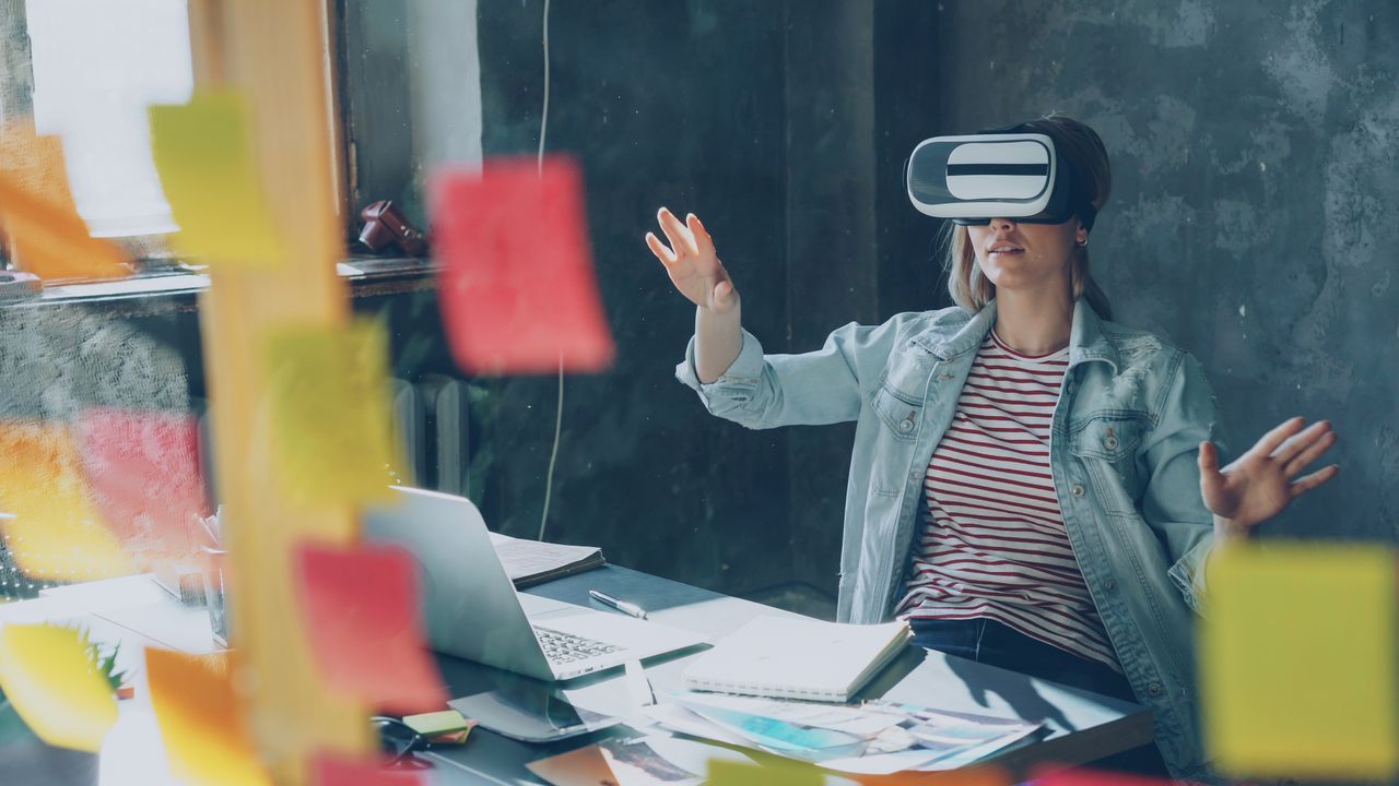 services-02 Woman in a creative workspace using a VR headset, surrounded by sticky notes and a laptop.